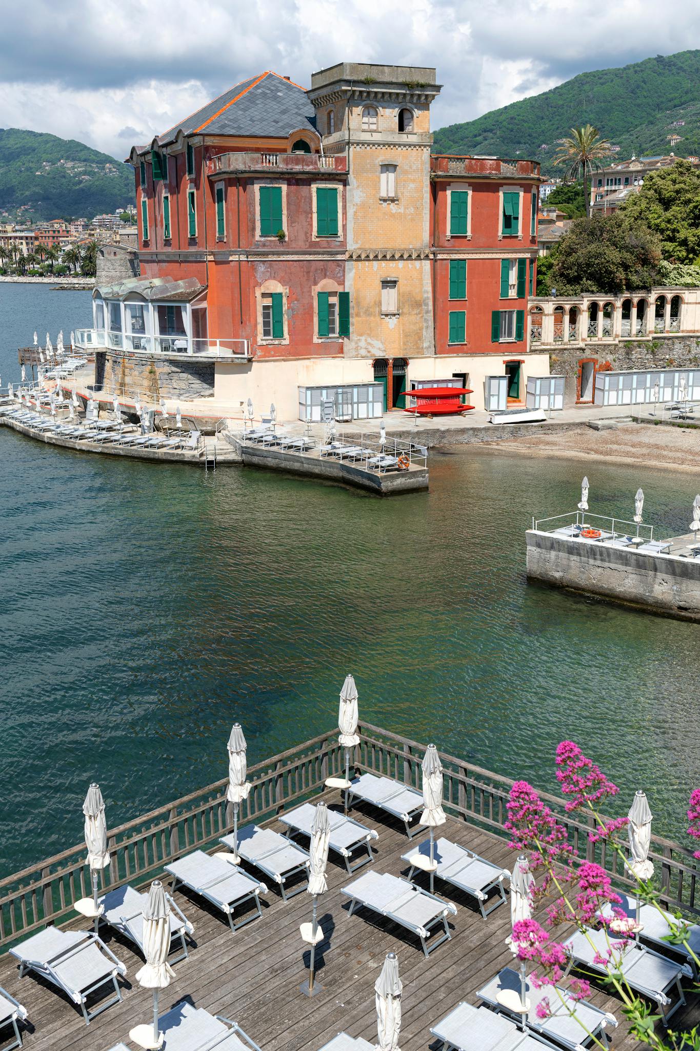 A picturesque scene of Rapallo, Liguria, featuring a historic building by the water and lounging chairs.
